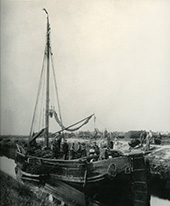 Turf laden, circa 1900. Het laden en opstapelen (loegen) van turf aan boord was vrouwenarbeid. Mannen brachten de turf vanuit het veld naar het schip. Op deze foto wordt een grote turfpraam geladen. De schippersvrouw (met muts) staat bij de helmstok. Uit de foto blijkt duidelijk dat het laden van een turfschip veel menskracht vergde.