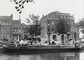 Beurtvaartdienst van Amsterdam naar Utrecht, circa 1920. Het beurtschip ligt afgemeerd aan een kade in Utrecht, met daarachter een bergplaats annex paardenstal (rechts van de straat), een winkel voor het halen en brengen van pakketten annex het kantoor (links van de straat), en de paard-en-wagen van de onderneming voor het ophalen en bezorgen van zware goederen.