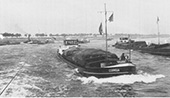 Druk binnenvaartverkeer in de scherpe Rijnbocht bij Stürzelberg in Duitsland, circa 1955. Op de voorgrond in de opvaart het kleine, geladen motorschip <em>Cornelia</em> dat aan de voormast de oploopvlag heeft gehesen, als teken dat het de voorgaande schepen wil passeren. Tegelijkertijd is aan de stuurhut de blauwe vlag uitgestoken, als teken dat het vaartuig de bakboordzijde (linkerzijde) van het vaarwater aanhoudt. Daardoor vaart het schip door de binnenbocht, waar de stroming van de rivier veel minder is dan in de buitenbocht. Aan de linkerzijde van de foto een afvarende sleep, voortgetrokken door een stoomsleepboot.